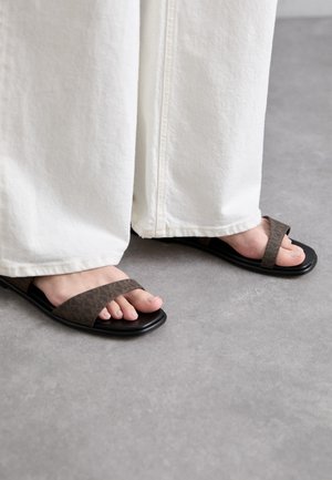 Sandale cu model maro, cu talpă neagră, având două curele late peste picior. Afișate împreună cu pantaloni largi albi.