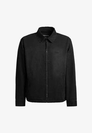 Giacca nera con cerniera, collo, maniche lunghe, tasche laterali e logo discreto sul petto sinistro, mostrata su sfondo bianco uniforme.