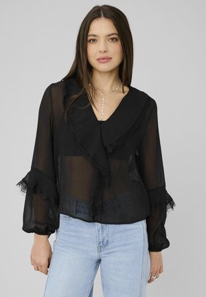 Femme portant une blouse noire transparente avec un col et des poignets à volants, un jean bleu clair et un délicat collier en argent, debout devant un fond uni.