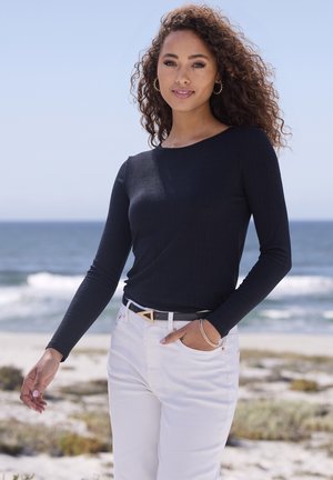 Schwarzes, geripptes Langarmoberteil kombiniert mit weißen Hosen, akzentuiert durch eine dreieckige Gürtelschnalle und ein Armband am Handgelenk. Meereskulisse.