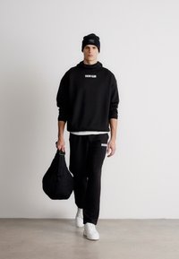 Čierna mikina a nohavice s logom, biele tričko, biele tenisky a čierna taška. Model má na sebe čiernu čiapku. Hladké textúry.