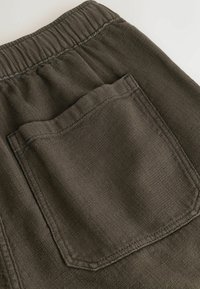 Gros plan sur un pantalon en tissu texturé vert olive foncé avec une taille élastique et une poche arrière rectangulaire avec des coutures visibles.