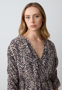 Leopardmuster-Kleid mit tiefem V-Ausschnitt, gerafftem Taillenbund, langen Ärmeln und weichem Stoff mit glatter Textur in Beige- und Schwarztönen.