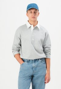 Jauns vīrietis ar zilu cepuri, pelēku garām piedurknēm polo krekls ar balto apkakli un gaiši zili džinsi, stāvot ar vienu roku kabatē.