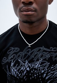 Valkoinen helminauha, jossa on pieni mietelause, makaamassa mustalla graafisella t-paidalla, jonka pinnassa on monimutkaisia hopeisia kuvioita ja rohkeaa tekstiä.