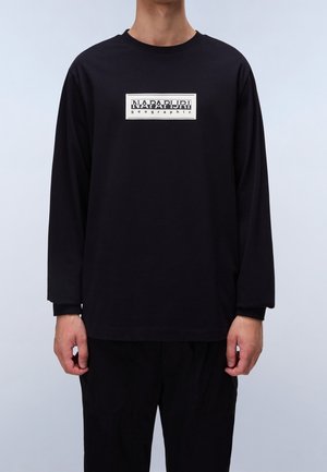 Mann trägt ein schwarzes Langarmshirt mit weißem rechteckigem "Napapijri geographic"-Logo in der Mitte der Brust, steht vor einem schlichten hellen Hintergrund.