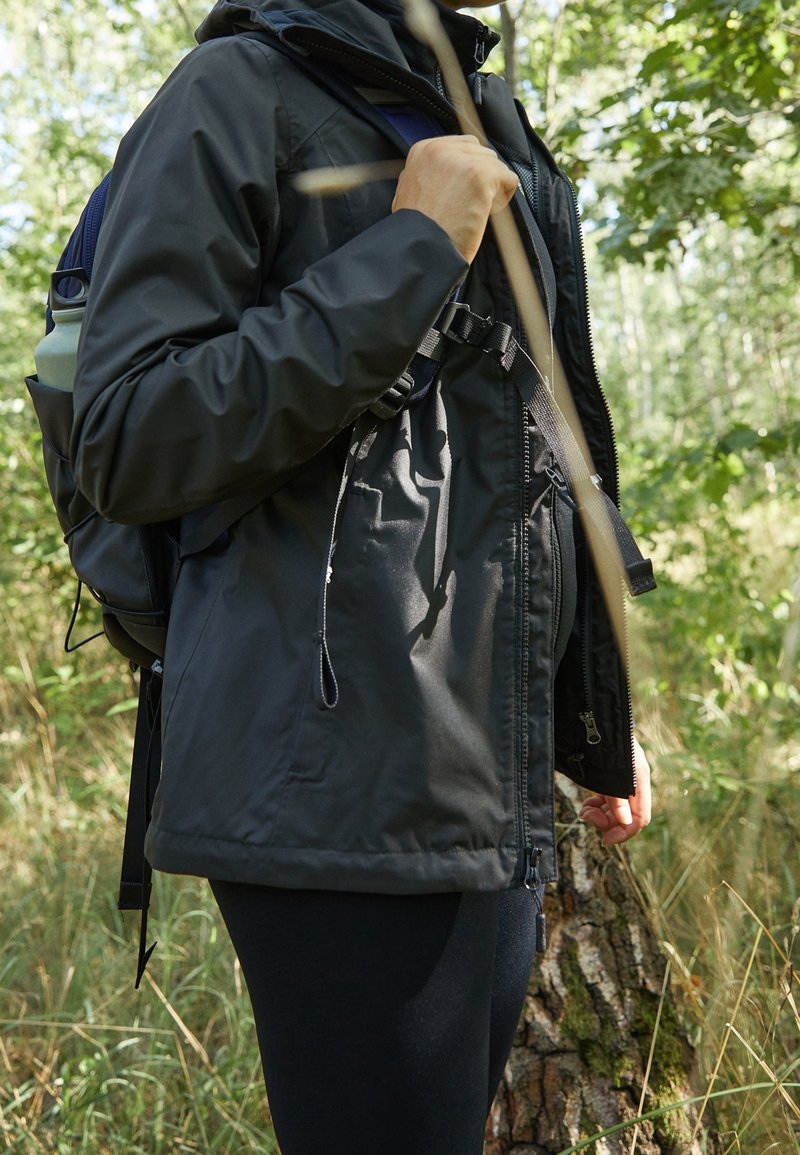 Chaqueta aislante negra con cremallera, capucha ajustable y bolsillos laterales, llevada sobre leggings negros, acompañada de una mochila en un entorno boscoso.