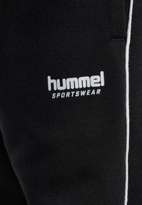 Czarny strój sportowy o miękkiej teksturze, zdobiony białym logo "hummel" oraz "SPORTSWEAR", podkreślony białym paskiem po bokach.