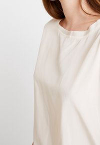 Blusa de un beige claro, hecha de un tejido suave. Presenta un escote redondo y mangas cortas. Textura suave con detalles de diseño mínimos.