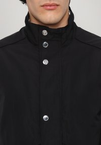 Black waterproof jacket with a high collar, front snap buttons, and smooth texture; features a minimalistic design with no visible patterns.