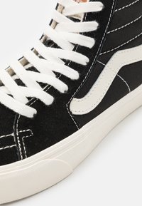 Zapatillas altas de canvas negras con cordones blancos, que presentan costuras en blanco y negro y un logo blanco característico en el lado.