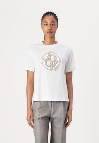 Weißes Baumwoll-T-Shirt mit einem runden Logo in einem beigen Muster in der Mitte. Kurze Ärmel und eine lässige Passform, kombiniert mit grauen karierten Hosen.