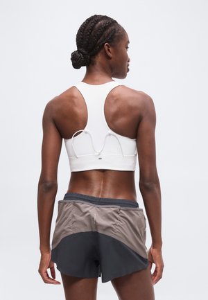 Mujer atlética con cabello trenzado, con un sujetador deportivo blanco y pantalones cortos de correr bicolor, de pie con la espalda hacia la cámara.