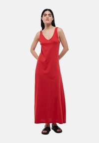 Robe maxi sans manches rouge avec un décolleté en V profond, fabriquée en tissu doux. Présente une coupe ample et une silhouette droite, associée à des sandales noires.