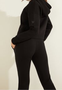 Sweat à capuche court noir avec capuche et poches latérales, associé à un pantalon noir ajusté. Présente un logo triangulaire sur la manche. Texture douce.