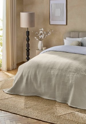 Bett mit beiger, strukturierter Decke und Kissen neben einer Holzlampe und Nachttisch mit Vase und Tasse in einem Schlafzimmer in neutralen Farben.