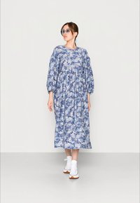 Mulher vestindo um vestido floral midi azul com mangas bufantes, tênis brancos e óculos de sol com tonalidade azul, posando contra um fundo claro e liso.