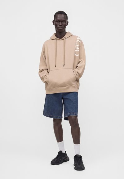 Homme debout portant un sweat à capuche beige avec un texte blanc, un short en jean bleu, des chaussettes blanches et des baskets noires sur un fond clair uni.
