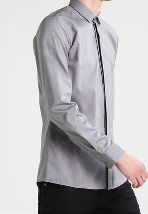 Mężczyzna ubrany w dopasowaną, długorękawową szarą koszulę z ukrytym listwą na guziki, stojący na tle jednolitego białego tła.