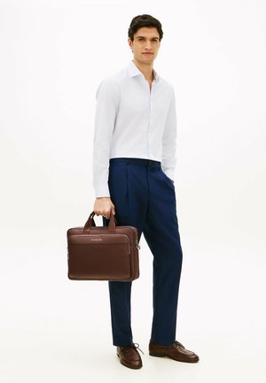 Hombre vistiendo camisa blanca y pantalones azul marino, sosteniendo un maletín de cuero marrón, de pie frente a un fondo blanco liso.