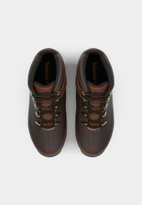 Las botas de senderismo de cuero marrón presentan un diseño de media caña, ojales metálicos y acentos en color beige con costuras en contraste. Branding de Timberland visible en el interior.