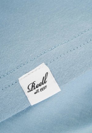 Tela azul clara con una textura suave. Presenta una etiqueta blanca con texto en negro que dice "Reell est. 1997", cosida en la costura.