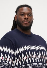 Tumsā zila adīta džemperis ar ģeometrisku baltu un sarkanu rakstu, apaļu kakla izgriezumu un strukturētu audumu.