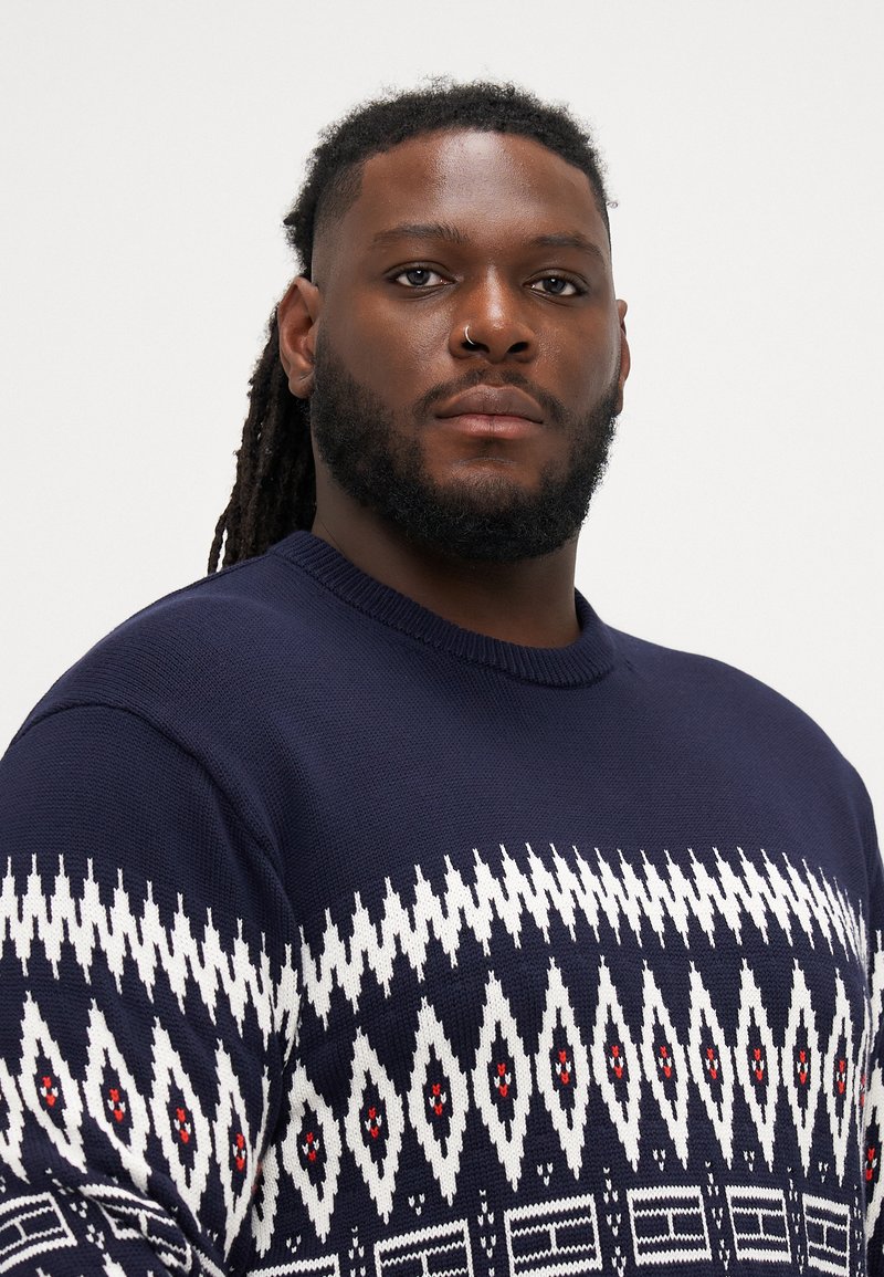 Navy blue knitted sweater featuring a geometric white and red pattern, round neckline, and textured fabric.