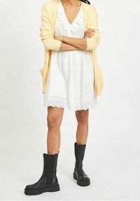 White dress with lace hem and collar detail, layered under a light yellow cardigan. Black knee-high boots with chunky soles.