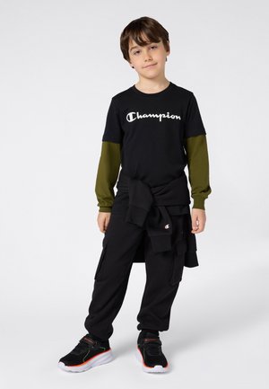 Jongen in een zwart Champion T-shirt met daaronder een olijfgroene lange mouw, zwarte cargo-broek, zwarte sneakers en een zwart sweatshirt om zijn taille geknoopt.