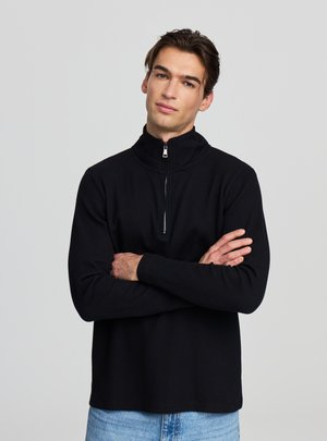 Maglione nero con zip, maniche lunghe e motivo a trama. Il colletto è rialzato e il tessuto è traspirante. Abbinato a jeans azzurri.