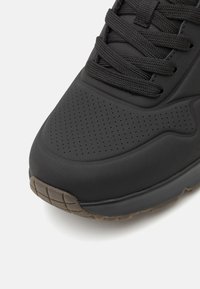 Sneaker negru cu un superior neted, perforat, vârf rotund și talpă din cauciuc texturat cu un model de grilaj. Sireturi sunt negre.