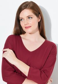 Vrouw met lichte huid en bruin haar, gekleed in een rode V-hals shirt, die naar de zijkant kijkt met de armen gekruist tegen een witte achtergrond.