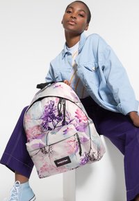 Blommönstrad ryggsäck, vit med lila och rosa accenter. Har en framficka och svart detaljer. Modellen bär en ljusblå jacka.