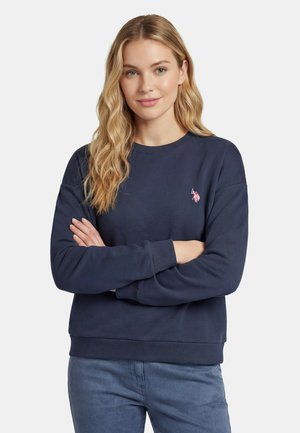 Woman with long blonde hair wearing a navy sweatshirt with a small pink embroidered logo, arms crossed, standing against a plain background.
