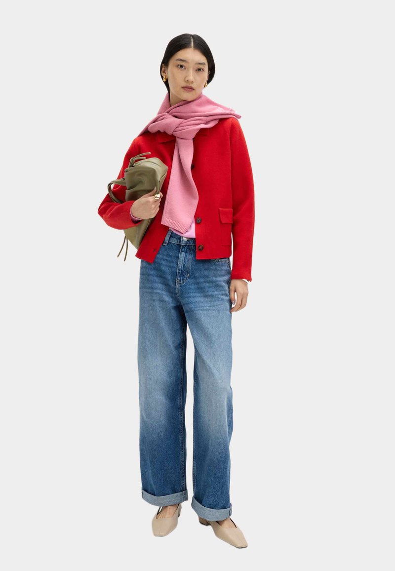 Giacca di lana rossa con bottoni, sciarpa rosa, jeans a gamba larga azzurri chiari e scarpe beige. Modella con borsa verde in mano, in piedi davanti a uno sfondo neutro.