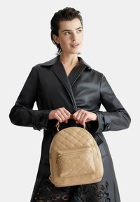 Zaino beige trapuntato con una tasca anteriore con zip, hardware dorato e una forma arrotondata, tenuto da una persona in un trench in pelle nera.
