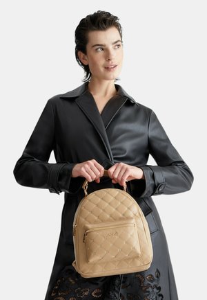 Zaino beige trapuntato con una tasca anteriore con zip, hardware dorato e una forma arrotondata, tenuto da una persona in un trench in pelle nera.