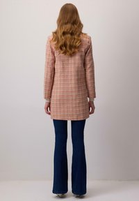 Kockovaný plášť v ružových a oranžových odtieňoch, s rovnými líniami, dlhými rukávmi a prispôsobenou siluetou, skombinovaný s rozšírenými tmavými džínsami.