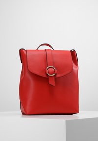 Zaino in pelle rossa con un flap anteriore, fissato da una cinghia con una fibbia circolare argentata, posato su una superficie bianca contro uno sfondo grigio.