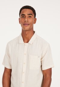 Korte mouwen, collared shirt in roomkleur met tan verticale strepen. Beschikt over een borstzak met een label en knoopsluitingen. Lichtgewicht stof.