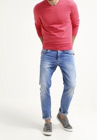 Maglione rosso a scollo a V, jeans azzurri chiari con effetto sbiadito e scarpe da ginnastica grigie. Tutti i capi sono casual e ben sagomati, con dettagli di cucitura visibili.