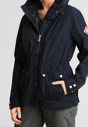 Kobieta ubrana w granatową kurtkę na zamek z wysokim kołnierzem i kieszeniami z patkami na napy, w połączeniu z szarymi spodniami, z ręką w kieszeni.