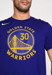 Camiseta de algodón púrpura con un gráfico redondo que presenta un diseño de puente, "30", y el texto "GOLDEN STATE WARRIORS" en amarillo. Logo de Nike en blanco.