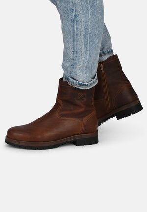 Botas de tobillo de cuero marrón con costuras claras, ligero brillo y suela de goma. Presentan una cremallera lateral y puntera redonda. Llevadas con jeans de color azul claro.