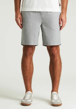 Homme portant un short gris clair longueur genou, des baskets blanches et une chemise blanche, debout les mains détendues le long du corps.