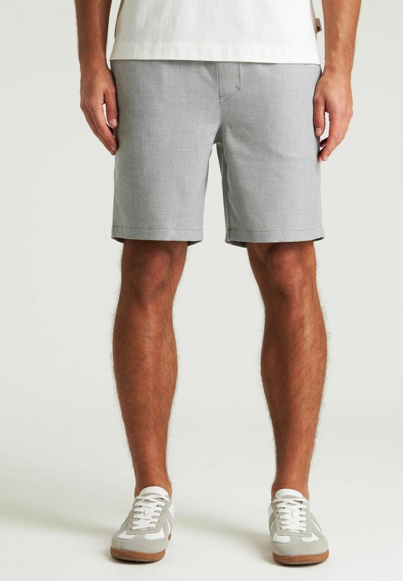 Homme portant un short gris clair longueur genou, des baskets blanches et une chemise blanche, debout les mains détendues le long du corps.