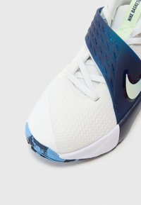 Hvid atletiksko med et tekstureret design, der har en mørkeblå rem og lyseblå detaljer på sålen. Nike-logoet er fremtrædende vist.