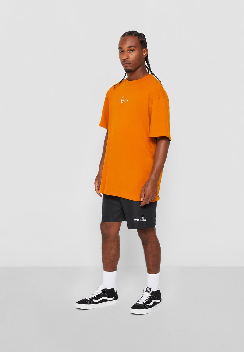 Tricou oversized portocaliu cu un logo alb, pantaloni scurți negri cu un logo, șosete albe și adidași negri. Material fin, croi lejer.