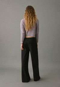 Maglione in stile uncinetto di colore lavanda con maniche lunghe, abbinato a pantaloni a gamba larga neri a vita alta. I capelli sono lunghi e mossi, visti da dietro.